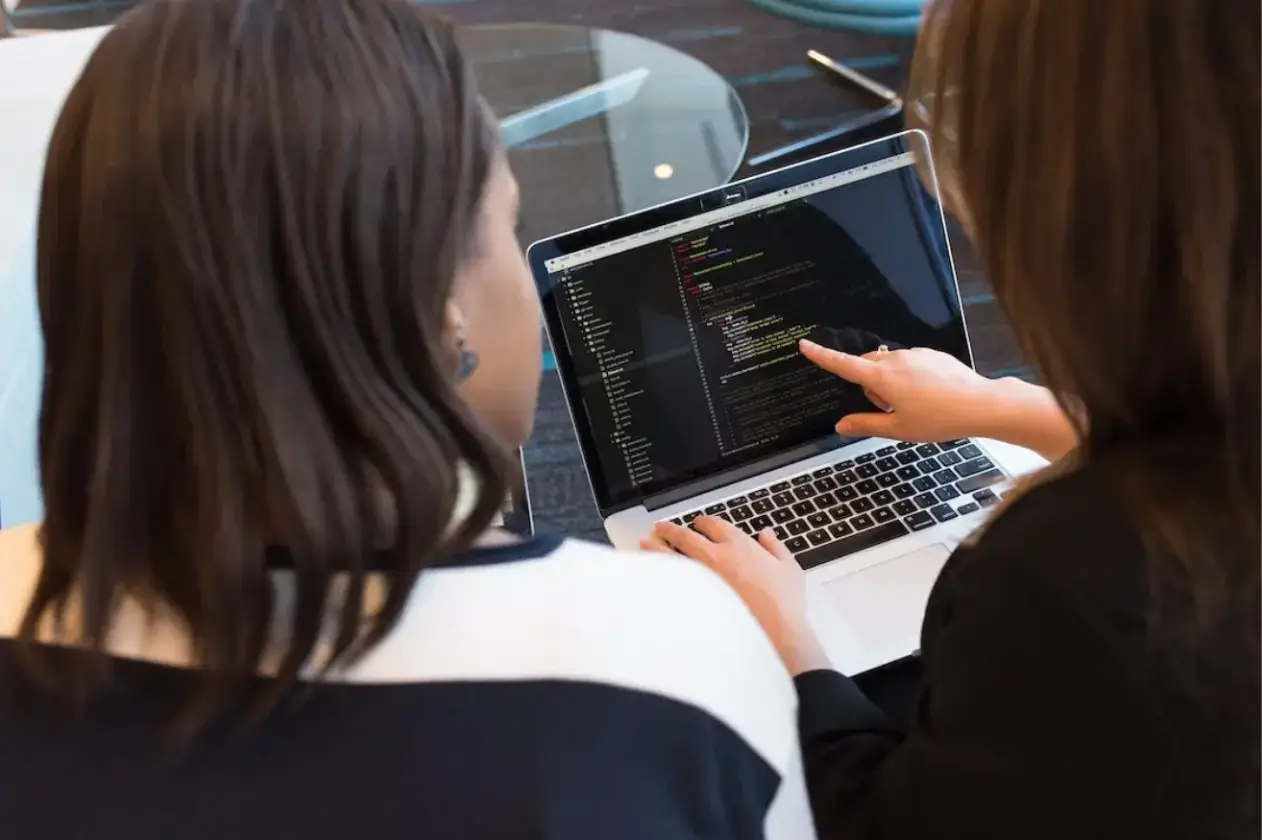 two-women-sitting-in-an-offic- environment-looking-at-a-laptop-displaying-programming-code